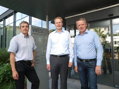 Drei Männer in Business-Kleidung stehen vor dem Eingang eines modernen Firmengebäudes mit WESCO-Firmenschild im Hintergrund