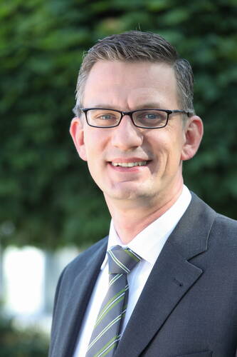 Lächelnder Mann mit Brille im Anzug und gestreifter Krawatte steht im Freien vor unscharfem grünen Hintergrund
