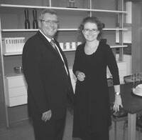 Zwei elegant gekleidete Personen stehen lächelnd in einem modern eingerichteten Büro mit Regalen und Tisch im Hintergrund