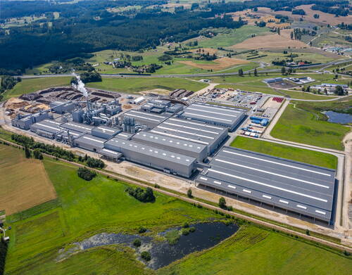 Großer Industriekomplex mit mehreren langgestreckten Hallen und rauchendem Schornstein, umgeben von Feldern, Wiesen und kleinen Teichen in einer ländlichen Landschaft aus der Luft gesehen
