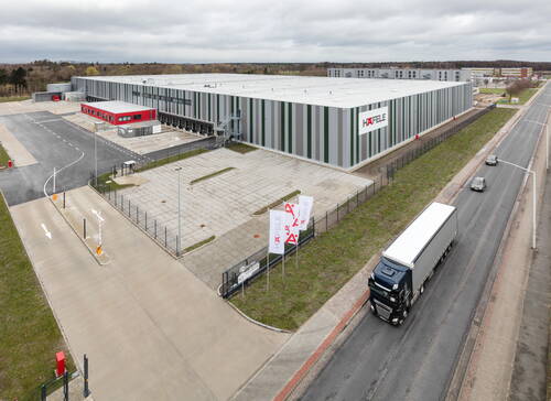 Großes, modernes Logistikzentrum mit grau-grün gestreifter Fassade und Häfele-Logo an einer Zufahrtsstraße, vor dem ein Lastwagen vorbeifährt.