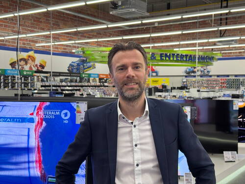 Mittelalter Mann im Business-Anzug lächelt in einer Elektronikfachmarkt‑Abteilung vor ausgestellten Fernsehern und Regalen mit Spielen und Unterhaltungsmedien