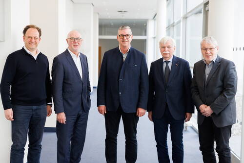 Fünf Männer mittleren bis höheren Alters stehen lächelnd nebeneinander in einem hellen Flur mit großen Fensterfronten und tragen überwiegend Anzug oder Sakko.