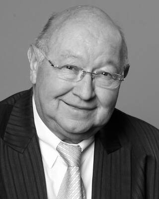 Älterer Mann mit Brille und Glatze lächelt freundlich in die Kamera und trägt Anzug mit Krawatte in einem schwarz-weißen Porträt.