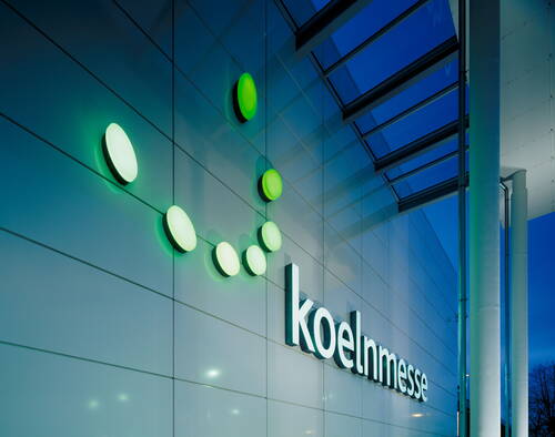 Leuchtende grüne Kreispunkte und der Schriftzug „koelnmesse“ an der modernen Glasfassade eines Messegebäudes in der Abenddämmerung