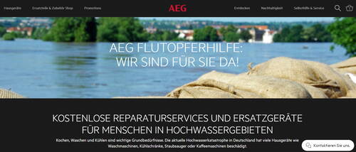 AEG informiert über kostenlose Reparaturservices und Ersatzgeräte als Flutopferhilfe für Menschen in Hochwassergebieten vor dem Hintergrund eines überfluteten Flusses mit Sandsäcken am Ufer