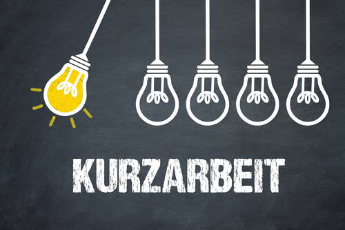 Leuchtende gelbe Glühbirne stößt gegen vier weiße Glühbirnen über dem Schriftzug „Kurzarbeit“ auf dunkler Tafelwand