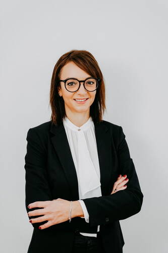 Junge Frau mit Brille und Business-Outfit steht mit verschränkten Armen vor hellem Hintergrund und lächelt selbstbewusst in die Kamera