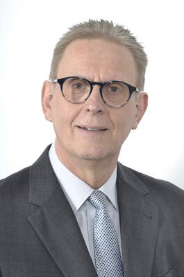 Älterer Mann mit kurzer, hellgrauer Frisur, dunkler Brille und grauem Anzug mit gemusterter Krawatte vor weißem Hintergrund