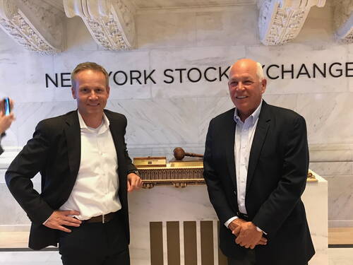 Zwei Geschäftsleute in Anzügen lächeln in der Halle der New York Stock Exchange vor einem Pult mit Richterhammer und Marmorsäulen im Hintergrund