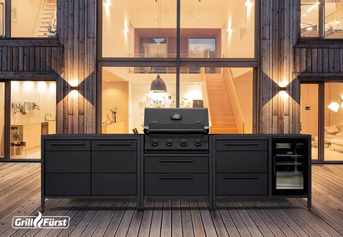Moderne Outdoor‑Küche mit schwarzem Gasgrill, Stauraum und Getränkekühlschrank auf einer Holzterrasse vor einem großzügig beleuchteten Haus mit Glasfront und Treppenaufgang