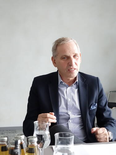 Älterer Mann im Anzug sitzt an einem Konferenztisch und spricht gestikulierend vor Wasser- und Getränkeflaschen.