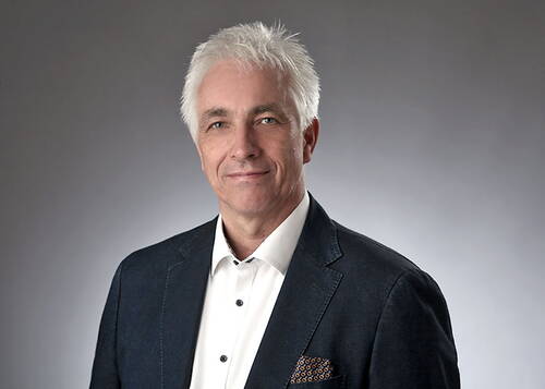 Älterer Mann mit weißem Haar im dunklen Sakko und weißem Hemd lächelt dezent vor neutral grauem Hintergrund in einem professionellen Porträt.