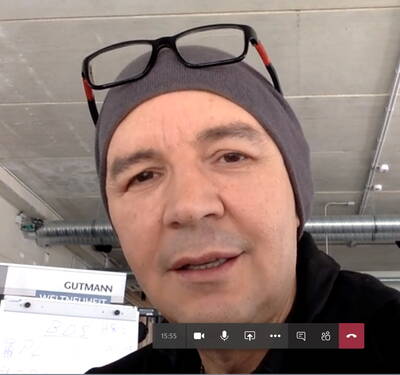 Mann mit grauer Mütze und hochgeschobener Brille spricht in einem Videocall in einem modernen Büro mit Sichtbetondecke und Whiteboard im Hintergrund.