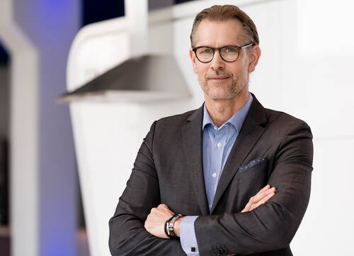 Geschäftsmann mittleren Alters mit Brille und verschränkten Armen steht selbstbewusst im modernen, hellen Büro.