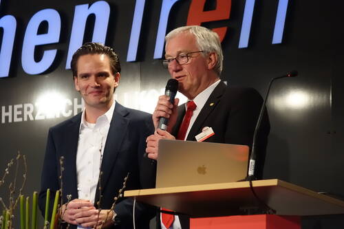 Zwei Männer im Anzug stehen auf einer Bühne, einer spricht mit Mikrofon neben einem Laptop am Rednerpult, während der andere lächelnd zuhört.