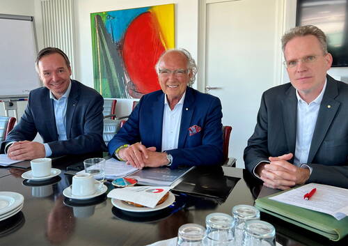 Drei Geschäftsmänner im Anzug sitzen lächelnd an einem Konferenztisch mit Unterlagen, Kaffeetassen und Gebäck vor einem großen, farbenfrohen abstrakten Gemälde im Besprechungsraum.