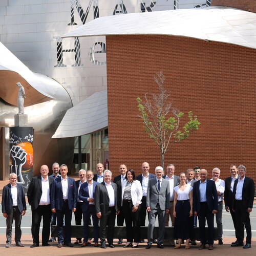 Gruppe von Geschäftsleuten in formeller Kleidung posiert im Freien vor moderner Architektur mit roter Backsteinfassade, gewellter Metallverkleidung, junger Baumallee und skulpturaler Stele mit Graffiti und erhobener Faust.