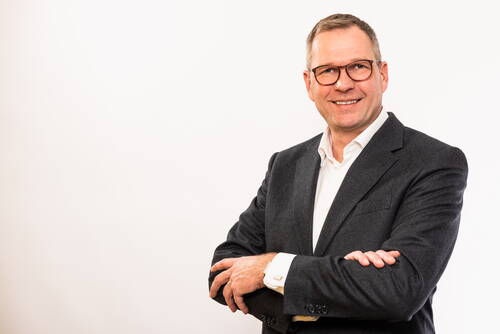 Geschäftsmann mittleren Alters mit Brille und grauem Anzug steht vor hellem Hintergrund und lächelt mit verschränkten Armen in die Kamera