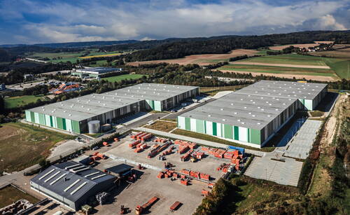 Großes Logistik- und Lagerzentrum mit mehreren grünen Hallen und weitläufigem Außenlager inmitten einer ländlichen Landschaft mit Feldern und Wäldern.