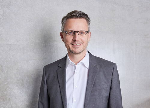 Mittelalterlicher Mann mit Brille und grauem Anzug lächelt vor einer hellgrauen Betonwand in die Kamera