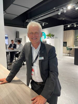 Älterer Mann im Anzug lehnt lächelnd an einem Holztresen in einer modernen Küchenausstellung, während im Hintergrund mehrere Personen ein Gespräch führen