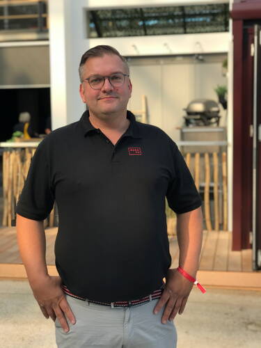 Mittelalterlicher Mann mit Brille und schwarzem Poloshirt steht entspannt im Freien vor einer modernen Terrasse mit Grill im Hintergrund