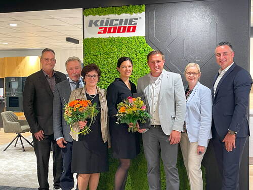 Sieben festlich gekleidete Personen posieren lächelnd im Küchenstudio vor einer grünen Mooswand mit dem Logo „Küche 3000“, zwei von ihnen halten bunte Blumensträuße.