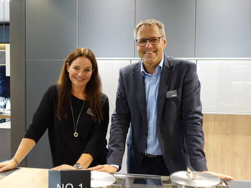 Zwei freundlich lächelnde Mitarbeitende in Businesskleidung stehen gemeinsam an einer modernen Kücheninsel in einer Ausstellungsküche.