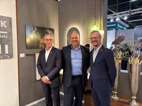 Drei lächelnde Männer in Anzügen posieren gemeinsam auf einer modernen Messeausstellung mit Wandbild eines Hirsches, dekorativen Leuchten und stilvollen Vasen im Hintergrund.