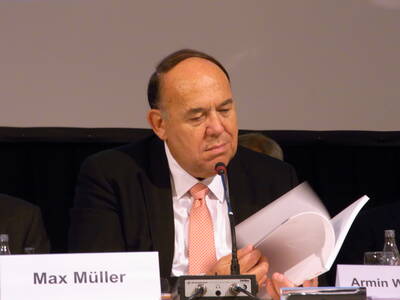 Älterer Mann im Anzug blättert konzentriert durch Unterlagen und spricht in ein Mikrofon bei einer Konferenz auf einem Podium.