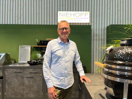 Mittelalter Mann mit Brille steht lächelnd in moderner Outdoor-Küche vor grünem Messestand mit Niehoff-Garden-Logo und großem schwarzen Keramikgrill im Vordergrund.