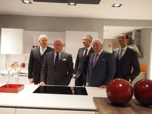 Fünf elegant gekleidete Männer im Anzug stehen lächelnd in einer modernen Ausstellungsküche mit Induktionskochfeld und großen roten Deko-Kugeln auf der Arbeitsplatte.