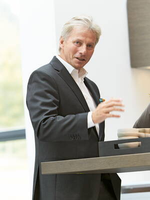 Älterer Mann im Anzug spricht gestikulierend an einem Stehtisch in einem hellen, modernen Raum