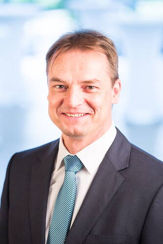 Lächelnder Mann im Anzug mit blau gestreifter Krawatte vor unscharfem, hellblauem Hintergrund