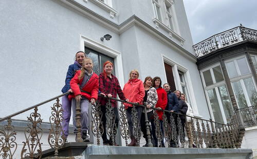 Mehrere Erwachsene und ein Kind stehen in bunter Winterkleidung lächelnd auf einem verzierten Balkongeländer vor einer hellen historischen Villa.