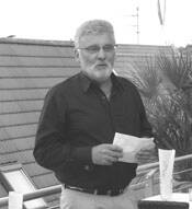 Älterer Mann mit grauem Bart hält Notizzettel in der Hand und spricht auf einer Terrasse im Freien