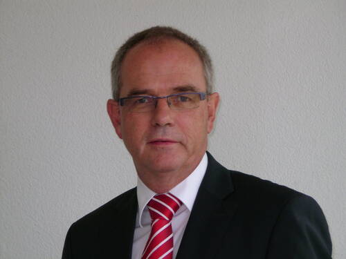 Älterer Mann mit Brille im dunklen Anzug und rot-weiß gestreifter Krawatte steht vor einer hellen Wand und blickt ernst in die Kamera.