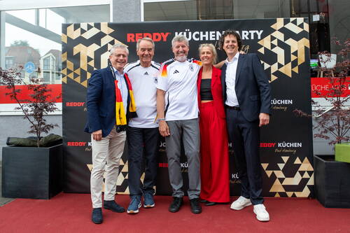 Fünf fröhlich lächelnde Menschen posieren im Business- und Fan-Outfit vor einer Reddy-Küchen-Eventwand auf rotem Teppich.