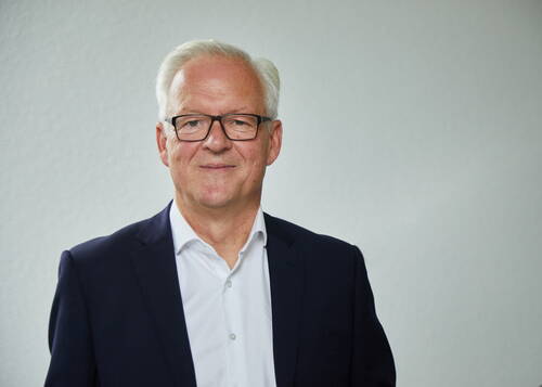 Älterer Mann mit grauen Haaren, Brille und dunklem Anzug blickt lächelnd vor neutral grauem Hintergrund in die Kamera.