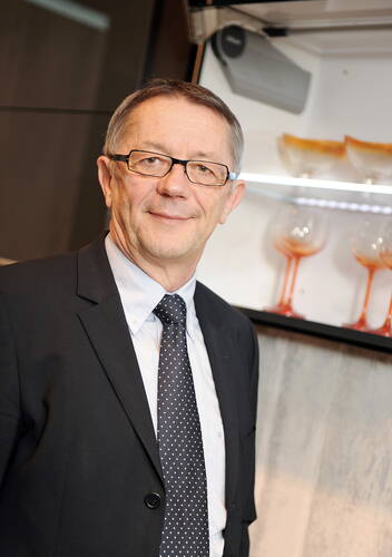 Älterer Mann im Anzug und mit Brille lächelt in einer modernen Küche vor einem offenen Geschirrschrank mit Gläsern