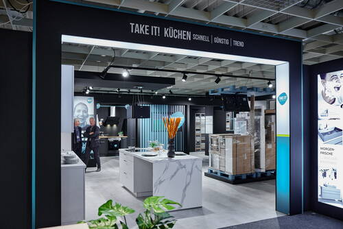 Modern gestaltete Küchenausstellung mit zentraler Kochinsel in Marmoroptik, verpackten Möbeln auf Paletten und zwei Männern im Anzug im Gespräch unter dem Schriftzug „TAKE IT! KÜCHEN – SCHNELL | GÜNSTIG | TREND“.