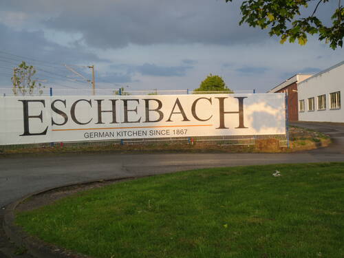 Großes Werbebanner mit der Aufschrift „Eschebach – German Kitchen since 1867“ an einem Zaun vor einem Industriegelände bei Abendlicht