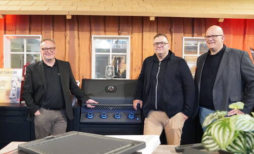 Drei Männer in Businesskleidung präsentieren in einer rustikalen Indoor-Kulisse einen modernen Gasgrill mit blau beleuchteten Drehknöpfen.