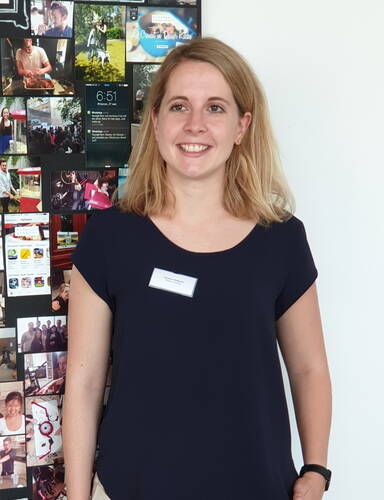 Junge Frau mit Namensschild lächelt vor einer Collage aus bunten Fotos an einer weißen Wand