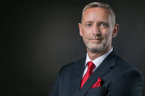 Reifer Geschäftsmann im dunklen Anzug mit roter Krawatte und Einstecktuch vor dunklem Hintergrund, der selbstbewusst in die Kamera blickt