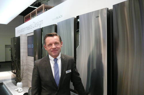 Mann im Anzug steht lächelnd vor einer Reihe moderner Edelstahl-Kühlschränke auf einer Design- und Technikmesse
