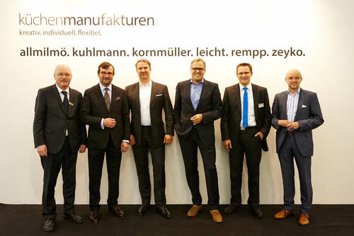 Sechs Geschäftsleute in Anzügen stehen lächelnd vor einer weißen Messewand mit dem Schriftzug „küchenmanufakturen“ und den Markennamen allmilmö, kuhlmann, kornmüller, leicht, rempp und zeyko.