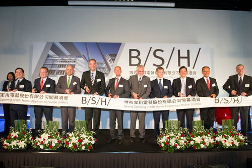 Geschäftsleute schneiden gemeinsam ein langes Band bei der feierlichen Eröffnung einer BSH-Niederlassung auf einer Bühne mit Blumenarrangements.