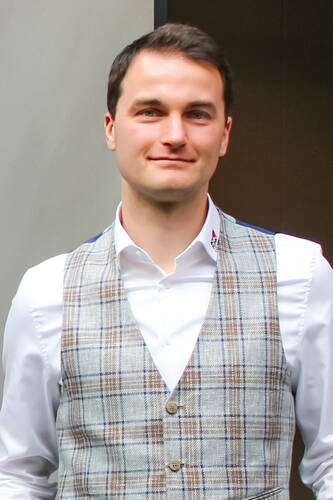 Junger Mann mit kurz geschnittenem dunklem Haar trägt ein weißes Hemd und eine hellgraue karierte Weste und blickt lächelnd in die Kamera.